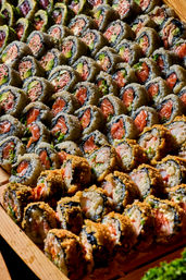 Close-up of a wooden platter piled with assorted sushi rolls — salmon and avocado maki, spicy crab and crispy tempura-style rolls neatly arranged