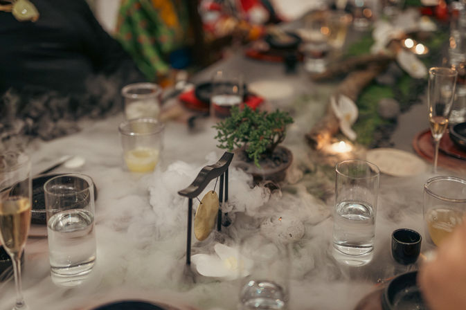 Festive dining table centerpiece with low-lying dry ice fog, small tabletop gong, miniature bonsai, scattered glassware and champagne flutes