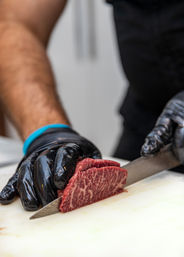 Gloved hands expertly slicing richly marbled raw steak with a sharp chef's knife on a white cutting board in a professional kitchen