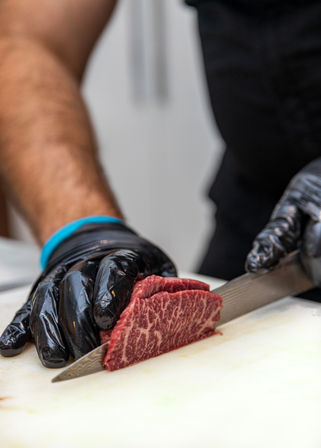 Gloved hands expertly slicing richly marbled raw steak with a sharp chef's knife on a white cutting board in a professional kitchen