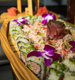 Wooden sushi boat with crab-topped rolls and avocado maki, a rose-shaped sashimi centerpiece and bright purple orchid garnishes