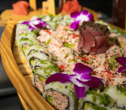 Wooden sushi boat with crab-topped rolls and avocado maki, a rose-shaped sashimi centerpiece and bright purple orchid garnishes