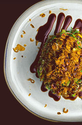 Overhead close-up of a gourmet appetizer: chopped greens and scallions topped with crunchy fried shallots, drizzled with a dark soy-style glaze on a white ceramic plate — modern restaurant plating.