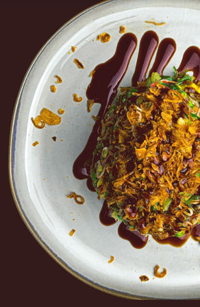 Overhead close-up of a gourmet appetizer: chopped greens and scallions topped with crunchy fried shallots, drizzled with a dark soy-style glaze on a white ceramic plate — modern restaurant plating.