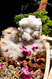 Japanese-style sushi boat platter with salmon and crispy rolls, glossy sauce, purple orchid flowers, curly kale garnish and dramatic dry-ice smoke pouring from a black bowl — elegant restaurant-style presentation.