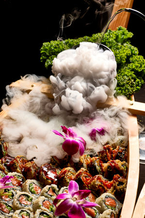 Japanese-style sushi boat platter with salmon and crispy rolls, glossy sauce, purple orchid flowers, curly kale garnish and dramatic dry-ice smoke pouring from a black bowl — elegant restaurant-style presentation.