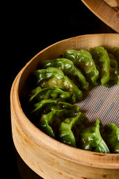 Vibrant green steamed dumplings with glossy pleated wrappers arranged in a round bamboo steamer basket on a black background.