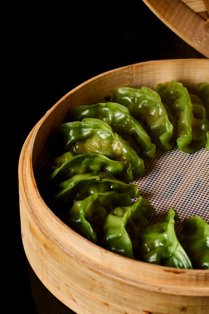 Vibrant green steamed dumplings with glossy pleated wrappers arranged in a round bamboo steamer basket on a black background.