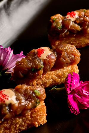 Close-up gourmet appetizer: diced seafood tartare with creamy sauce and red roe atop crispy crostini, garnished with bright pink edible flowers on a black plate.