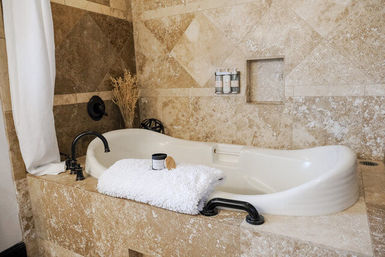 Spa-like beige stone-tiled bathroom with a built-in white soaking tub, oil-rubbed black fixtures, folded white towel and bath products