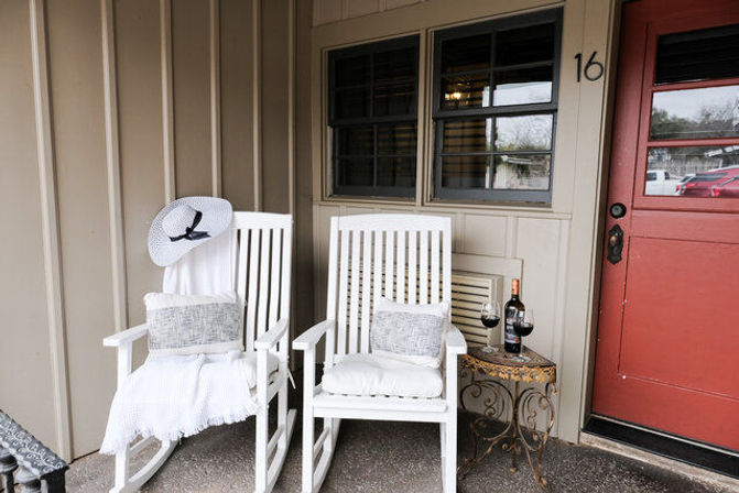Cozy front porch with two white wooden rocking chairs with cushions and a sun hat, ornate side table holding a bottle of red wine and two glasses, beside a red entry door and paneled siding.