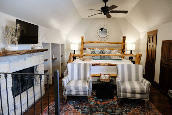 Cozy rustic bedroom in a lodge-style cabin with a log wood bed, patterned rug, two striped armchairs facing a stone fireplace with wooden mantel, round wall mirror, bedside lamps and ceiling fan