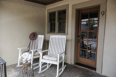 Cozy front porch with two white rocking chairs and cushions, a draped blanket and hat, an ornate iron side table with two mugs, and a wood‑framed glass entry door