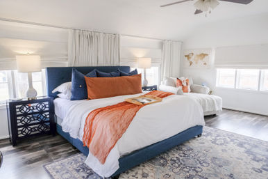 Bright modern master bedroom with navy upholstered king bed, white bedding, orange accent throw, patterned area rug, wood floors, and a cozy chaise by sunlit windows.