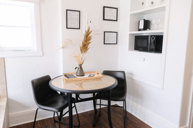 Sunny small apartment dining nook with round glass-topped table, two black chairs, wooden tray and vase of dried grasses, white built-in shelf with microwave and framed wall art