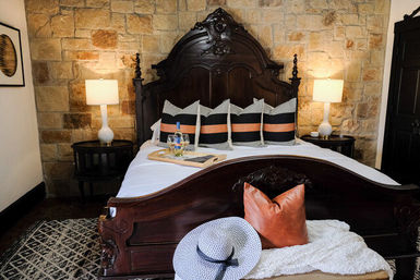 Ornate carved dark-wood bed with white linens and black-and-orange striped pillows set against a warm stone accent wall, matching bedside tables with lamps, a tray with wine on the bed, patterned rug and a straw sunhat on a bench.