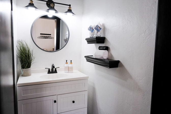 Chic modern white bathroom vanity and round mirror with black three-light sconce, black faucet, soap dispensers, potted grass and floating shelves holding a rolled towel labeled 'Makeup'.