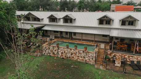 Aerial view of a rustic metal-roof lodge with dormer windows, an elevated stone-walled pool with potted plants, patios with lounge and dining seating, and trees on a grassy slope.