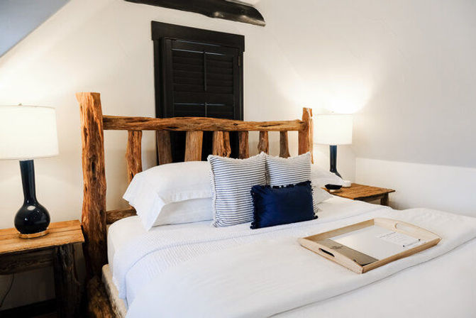 Cozy rustic loft bedroom with a log-frame bed, crisp white linens, striped and navy accent pillows, wooden bedside tables with black ceramic lamps, shuttered window and a serving tray on the bed.