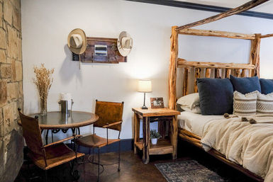 Cozy rustic cabin bedroom with a log four-poster bed, navy pillows and neutral bedding, wooden nightstand and lamp, round bistro table with leather chairs and a stone accent wall