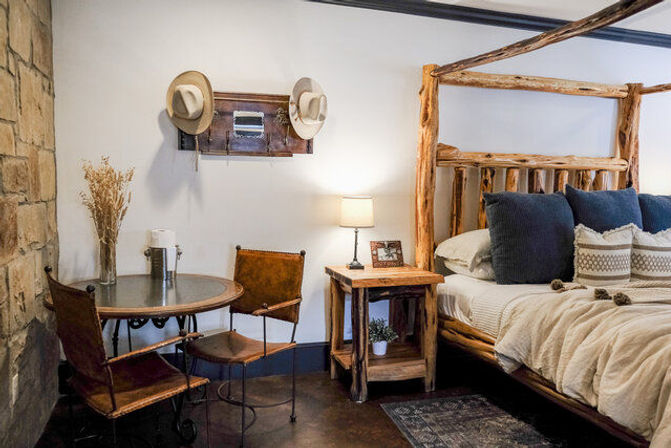 Cozy rustic cabin bedroom with a log four-poster bed, navy pillows and neutral bedding, wooden nightstand and lamp, round bistro table with leather chairs and a stone accent wall