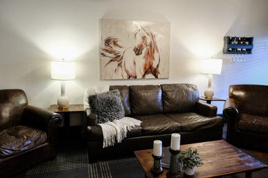 Cozy modern farmhouse living room interior — dark leather sofa and matching armchairs beneath a large horse painting, warm table lamps flanking the couch, wooden coffee table with candles and a small potted plant.