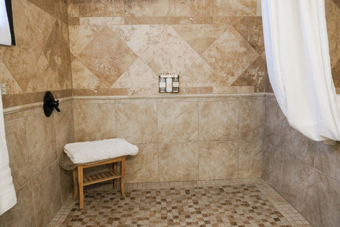 Beige stone-tiled walk-in shower with mosaic tile floor, small wooden bench with white towel, wall-mounted soap dispensers and white shower curtain