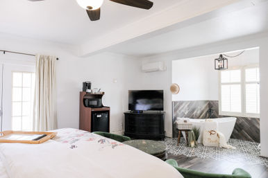 Bright modern guest suite with floral bedspread, ceiling fan, flat-screen TV, coffee station and mini fridge, open-plan ensuite with freestanding bathtub, patterned tile and wood floors, and shuttered window.