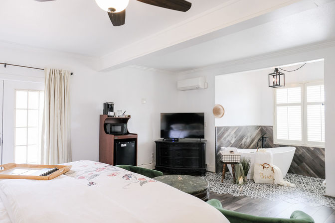 Bright modern guest suite with floral bedspread, ceiling fan, flat-screen TV, coffee station and mini fridge, open-plan ensuite with freestanding bathtub, patterned tile and wood floors, and shuttered window.