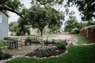 Shady suburban backyard patio with black metal dining set, rock-bordered herb garden, large oak tree, gravel driveway and wooden fence