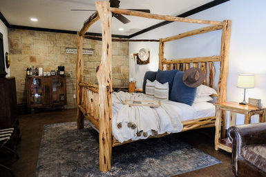 Cozy rustic lodge bedroom featuring a natural log four‑poster bed with white bedding and blue accent pillows, stone accent wall, wooden nightstand with lamp, vintage cabinet and leather chair on a patterned rug.
