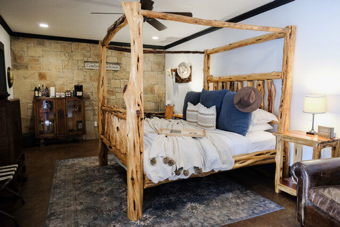 Cozy rustic lodge bedroom featuring a natural log four‑poster bed with white bedding and blue accent pillows, stone accent wall, wooden nightstand with lamp, vintage cabinet and leather chair on a patterned rug.