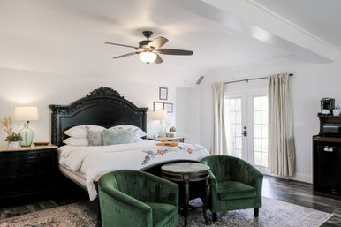 Bright cozy bedroom with ornate black carved headboard, king bed with white floral bedding, two green velvet chairs and round table, ceiling fan, French doors with neutral curtains and bedside lamps