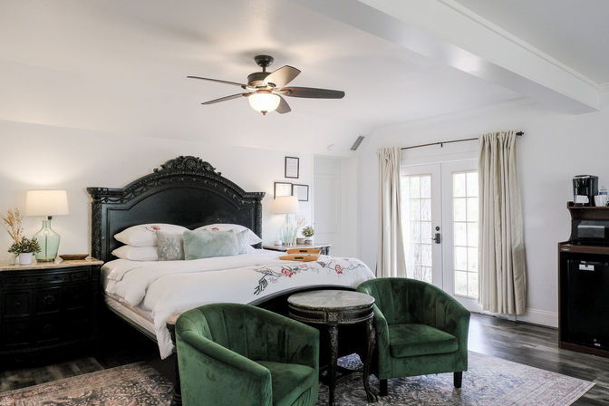 Bright cozy bedroom with ornate black carved headboard, king bed with white floral bedding, two green velvet chairs and round table, ceiling fan, French doors with neutral curtains and bedside lamps
