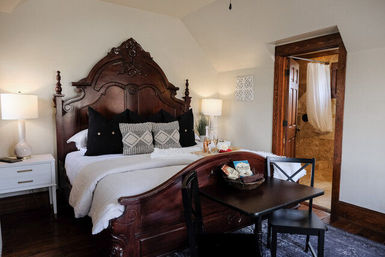 Ornate carved wooden bed with white linens and patterned pillows in a cozy boutique-style bedroom, bedside lamps and small table with chairs, open doorway to tiled ensuite bathroom