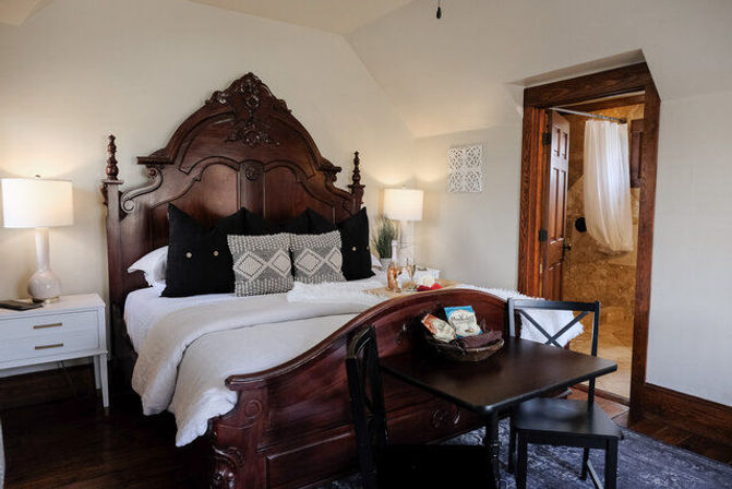 Ornate carved wooden bed with white linens and patterned pillows in a cozy boutique-style bedroom, bedside lamps and small table with chairs, open doorway to tiled ensuite bathroom