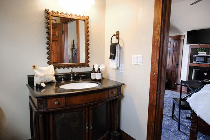 Cozy rustic en-suite bathroom vanity with carved wooden mirror, black stone countertop and oval sink, towel ring and toiletries on the counter, open doorway into an adjoining bedroom.