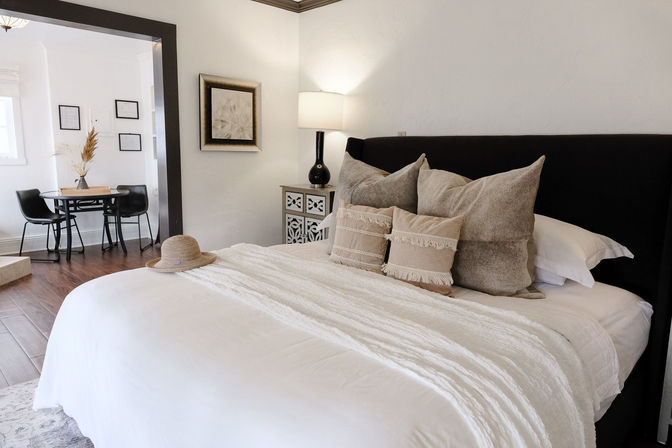 Cozy modern bedroom interior with white linens and textured beige throw pillows on a bed with a black headboard, a straw hat on the foot of the bed, bedside lamp on a decorative nightstand, and a small dining nook visible through an open doorway.