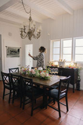 Sunlit farmhouse dining room with wooden table set for brunch, lush floral centerpieces and champagne flutes, vintage chandelier, terracotta tile floor, person holding a toasting glass by bright windows.