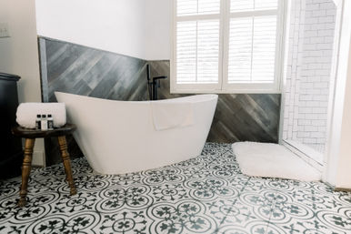 Bright spa-like farmhouse master bathroom with freestanding white tub, black-and-white patterned floor tiles, diagonal wood accent wall, subway-tiled walk-in shower, white shutters and plush bath mat.