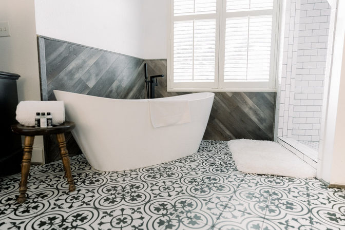 Bright spa-like farmhouse master bathroom with freestanding white tub, black-and-white patterned floor tiles, diagonal wood accent wall, subway-tiled walk-in shower, white shutters and plush bath mat.