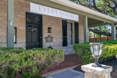 Quaint historic limestone house front with covered porch, black entry door and shuttered window, white rectangular sign above, manicured hedges and a lantern-style lamp on a stone post.