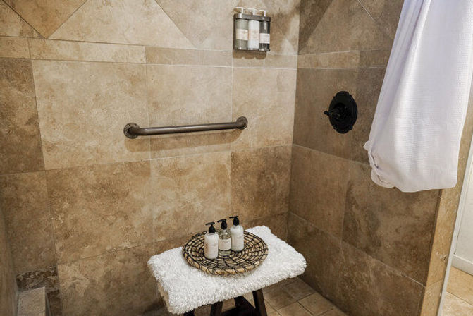 Spa-like beige stone-tile walk-in shower with safety grab bar, black shower control, hanging white towel, and a small stool topped with a fluffy cover, woven tray and three pump bottles.