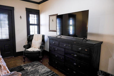 Cozy bedroom corner with a large flat-screen TV on a distressed black dresser, a leather armchair draped with a cream throw, shuttered windows, dark trim, and a textured area rug.
