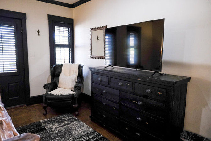 Cozy bedroom corner with a large flat-screen TV on a distressed black dresser, a leather armchair draped with a cream throw, shuttered windows, dark trim, and a textured area rug.