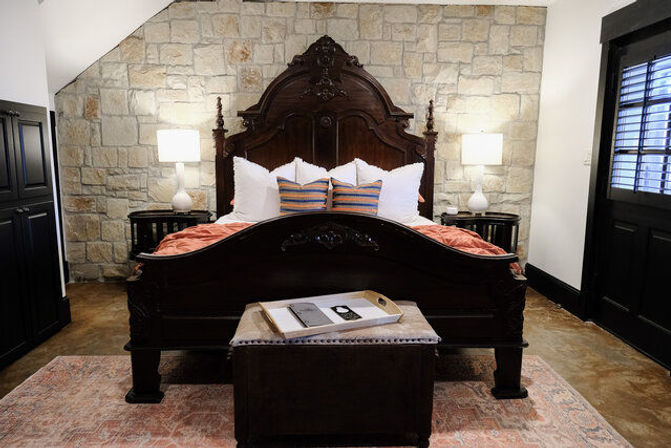 Rustic-chic bedroom with an ornate dark wood bed against a stone accent wall, striped throw pillows, bedside lamps, ottoman with tray, and shuttered window