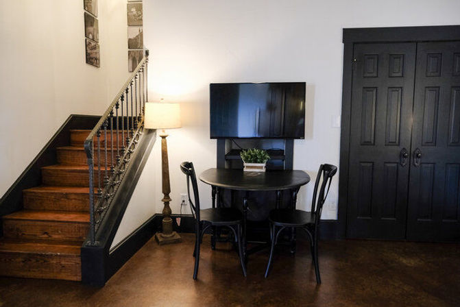 Interior living nook with wooden staircase, tall floor lamp, wall-mounted TV above a small round black table with two black chairs and a potted plant, beside dark double closet doors.