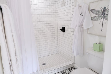 Bright modern bathroom with white subway-tile walk-in shower, black shower fixture, hex-tile shower floor, patterned floor tile, white robe and towels, dragonfly wall art