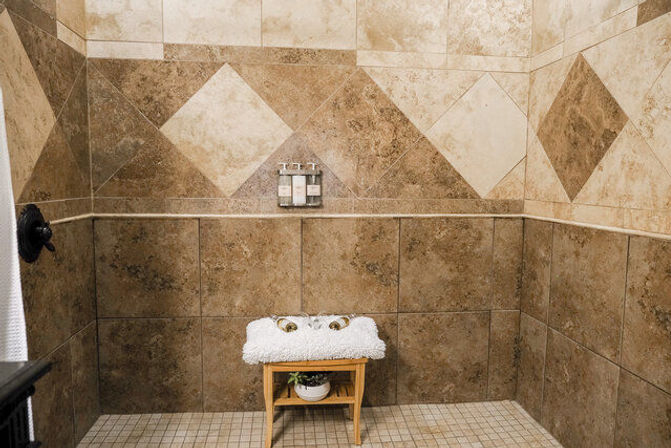 Spa-style walk-in shower with beige and brown stone tiles in a diamond pattern, wall-mounted soap dispensers, wooden stool topped with a fluffy white towel and a small potted plant on the lower shelf.