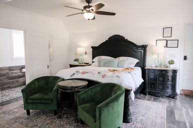 Bright, airy master bedroom with ornate black carved headboard, white floral-embroidered bedding, two green velvet accent chairs and round wooden side table, matching black nightstands with lamps, ceiling fan, patterned area rug and wood-look flooring — cozy, elegant home retreat.